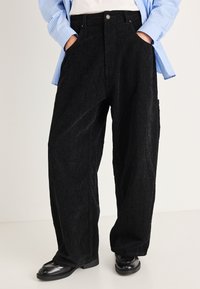 Fekete kordbársony bő szárú nadrág elülső zsebekkel és magas derékkal. Világoskék csíkos inggel és fekete cipővel párosítva.