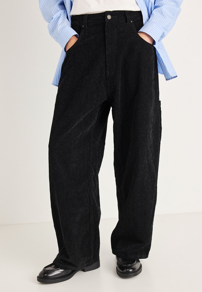 Fekete kordbársony bő szárú nadrág elülső zsebekkel és magas derékkal. Világoskék csíkos inggel és fekete cipővel párosítva.