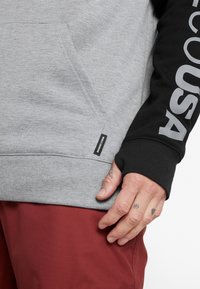 Felpa grigia con una grande manica nera che presenta testo bianco. Tasca a marsupio e polsini a coste. Etichetta visibile sull'orlo inferiore. Pantaloni rossi sotto.