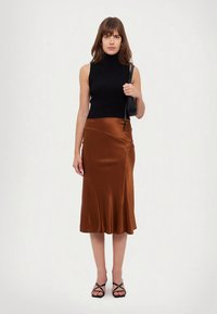 Kvinne iført en svart ermeløs poloskjorte, bronsefarget sateng midi-skjørt, svarte sandaler med remmer, bærende på en liten svart skulderveske.