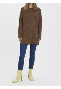 Brązowy, dziergany, oversize'owy sweter z długimi rękawami, zestawiony z niebieskimi dżinsami i żółtymi botkami na obcasie. Prosty fason i faktura.