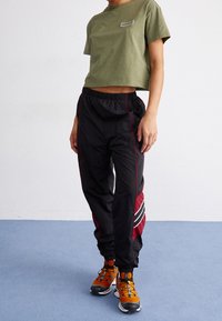 T-shirt court vert olive avec une étiquette, associé à un pantalon noir avec des rayures rouges et un design sur le côté. Des chaussures de sport orange complètent le look.