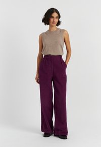 Tan mouwloze gebreide top met een geribbelde halslijn, gecombineerd met hoge taille, wijdvallende paarse corduroy broeken. Zwarte schoenen maken de look compleet.