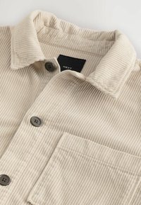 Beige corduroy shirt with a textured surface, featuring a fold-down collar, two buttons, and a single pocket. Label visible inside.