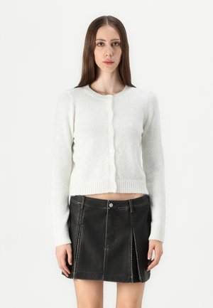 White knit cardigan with a round neckline, front buttons, and ribbed hem; worn over a black leather mini skirt with structured seams.