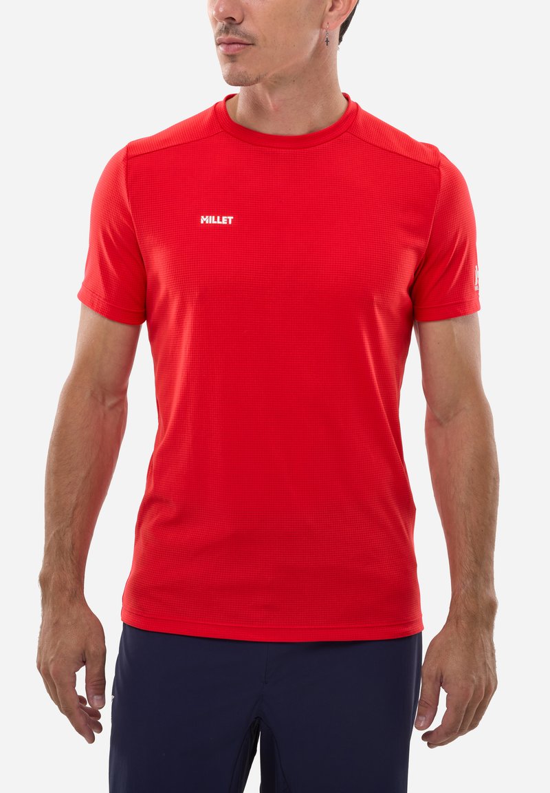 Homme portant un maillot de sport Millet à manches courtes rouge et un short foncé, debout devant un fond clair uni, regardant sur le côté.