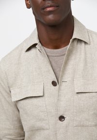 Camicia button-up beige chiaro realizzata in tessuto testurizzato, con due tasche sul petto e colletto. Sotto, è visibile una maglietta tinta unita color cammello.