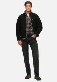 Giacca in pile nera con fodera beige, tasca con zip, indossata sopra una camicia a scacchi. Abbinata a jeans scuri e scarpe nere.