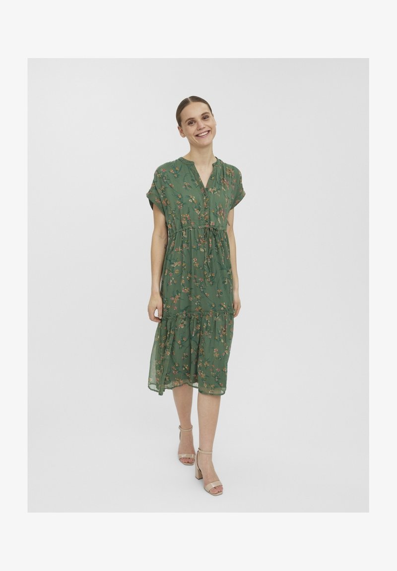 Femme souriante marchant, portant une robe midi verte à fleurs avec des manches courtes et des talons carrés beiges à bride cheville.