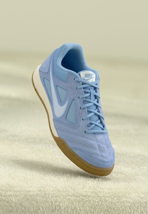 Light blue Nike sneaker with white swoosh and gum sole positioned on cream textured surface, angled to show front and side details.