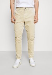 Pantalons en velours côtelé beige à coupe droite, avec poches avant et fermeture à un bouton, associés à des baskets blanches.