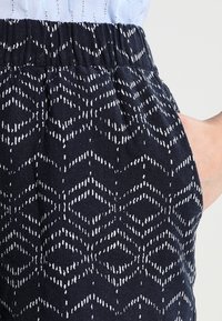 Kalhoty s námořnickým vzorem a strukturovaným tkaním, které mají bílý geometrický diamantový design a elastický pas. Viditelné otevřené boční kapsy.