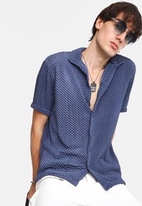 Camisa de manga corta azul marino con superficie texturizada, parte delantera abotonada y cuello extendido. Combinada con pantalones blancos y un collar en capas.