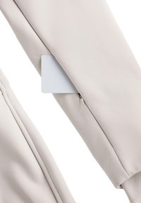 Jasnobeżowy rękaw kurtki na zamek błyskawiczny, wykonany z gładkiej, matowej tkaniny, z delikatnymi detalami szwów i pustą białą metką włożoną do kieszeni na zamek.