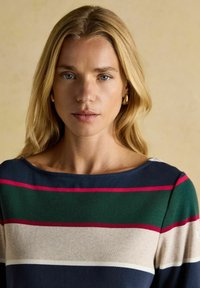 Blonde Frau mit blauen Augen, die goldene Creolen und einen mehrfarbigen gestreiften Pullover in Marineblau, Grün, Beige und Rot trägt.