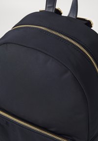 La mochila de tela negra presenta una textura suave y lisa, detalles de cremallera dorada y dos correas de cuero. Diseño compacto y redondeado con un bolsillo frontal.
