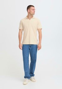 Polo beige avec col classique, associé à un jean en denim bleu. Le modèle se tient sur un fond neutre, portant des baskets crème.