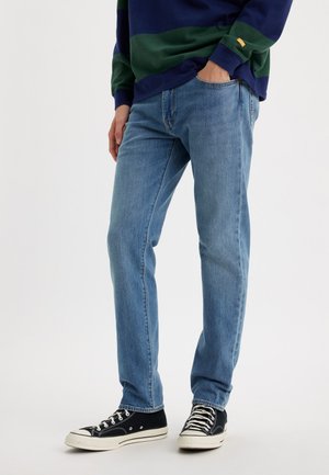 Blaue Jeans aus Denim mit geradem Schnitt, ausgestattet mit Fronttaschen, dezenten Stickdetails und kombiniert mit schwarz-weißen Canvas-Sneakern.