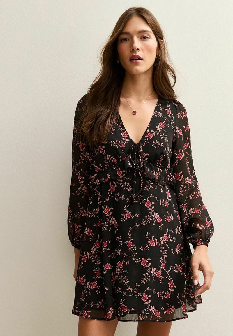 Vestido negro con flores que presenta un escote en V, detalles de volantes en el torso y mangas largas abullonadas. Patrones de pequeñas flores rosas y rojas a lo largo de toda la prenda.