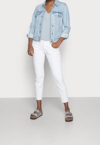 Frau in einer hellblauen Denimjacke, bluse in hellem Blau, weißen Cropped-Jeans und grauen Sandalen mit doppeltem Riemen, die mit einer Hand in der Tasche steht.