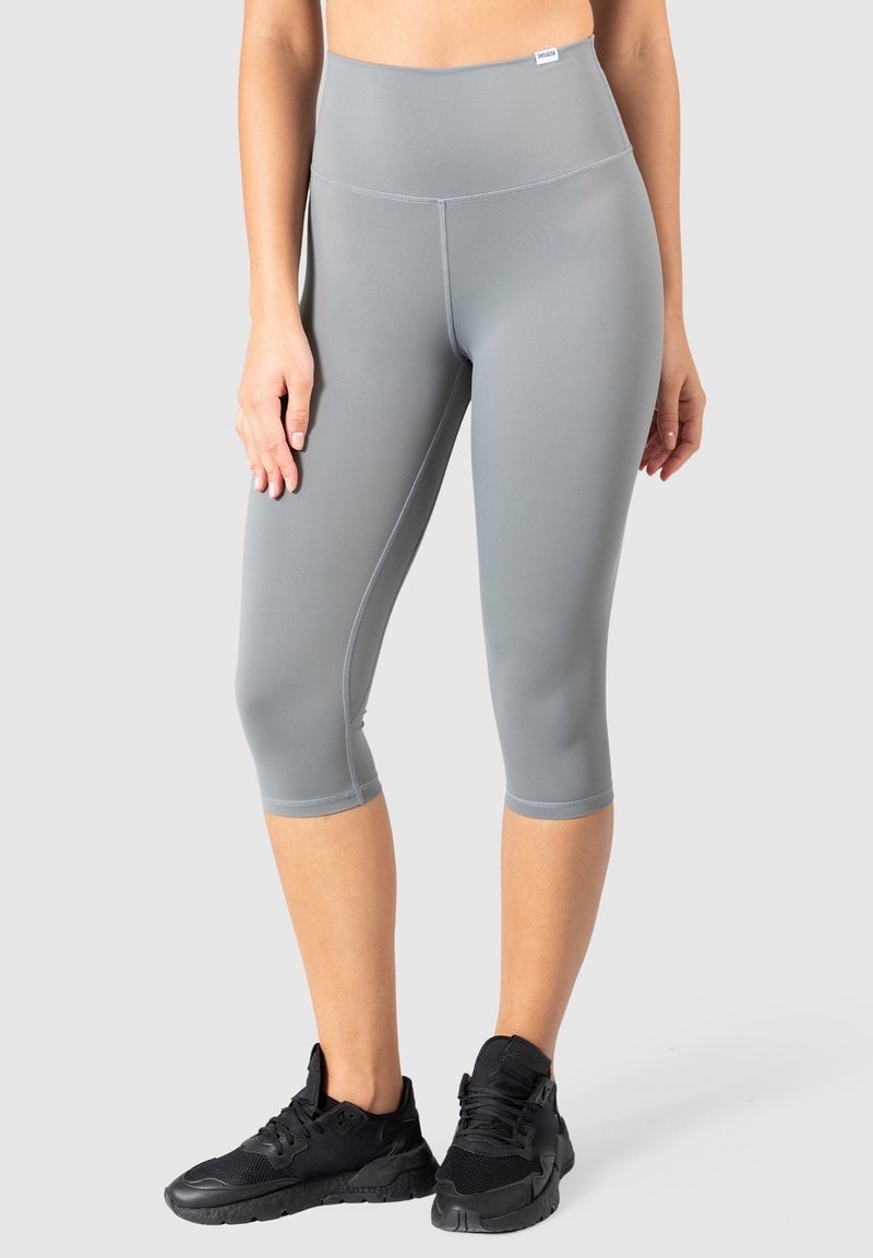 Femme portant des leggings gris courts taille haute et des chaussures de sport noires, debout devant un fond gris clair uni.