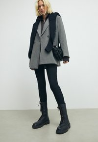Cappotto grigio doppiopetto con un maglione nero lavorato a maglia sulle spalle, abbinato a leggings neri e stivaletti combat neri con lacci.