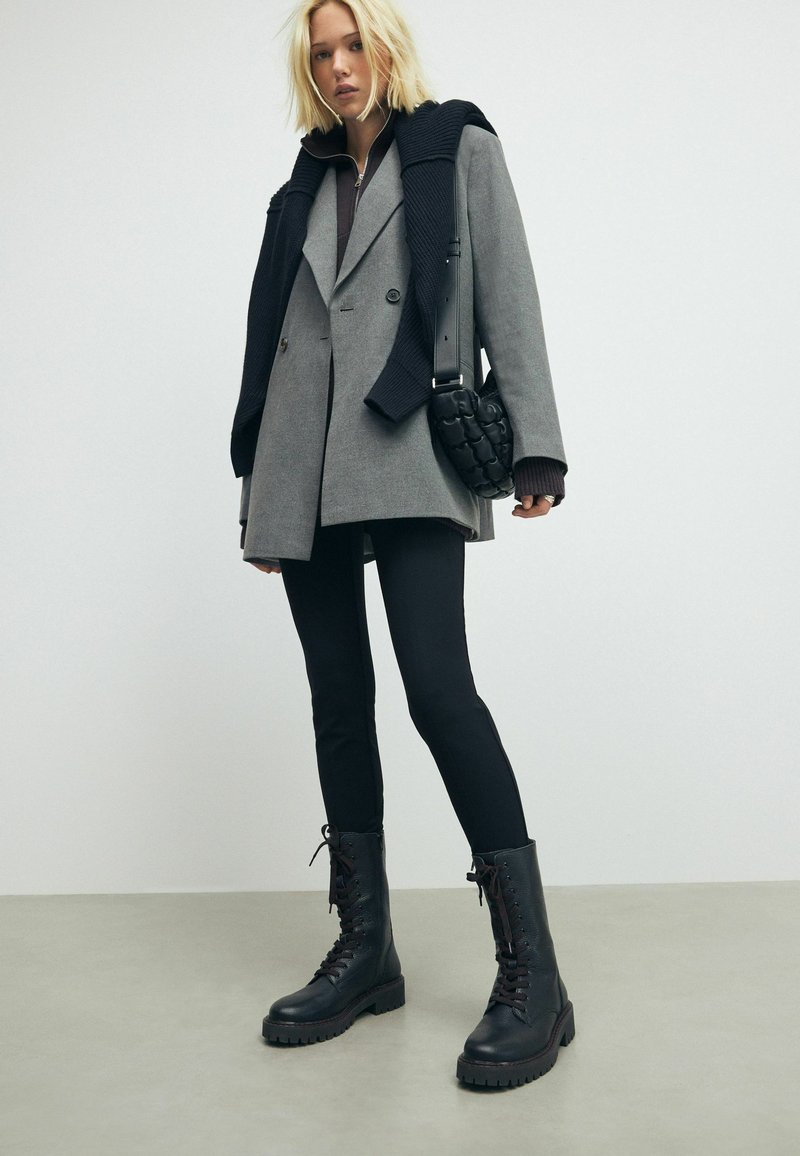 Cappotto grigio doppiopetto con un maglione nero lavorato a maglia sulle spalle, abbinato a leggings neri e stivaletti combat neri con lacci.