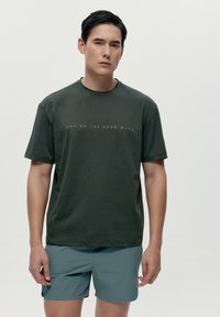 Temno zelena bombažna majica s kratkimi rokavi in okroglim ovratnikom, ki ima diskreten bel napis "HOP ON THE BORN WAVE" čez prsi.