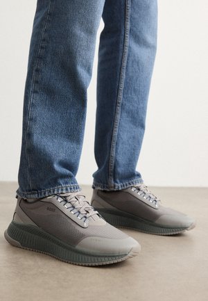 Persoon die een blauwe spijkerbroek en grijze BOSS-sneakers draagt, staand op een gladde vloer tegen een effen witte achtergrond.