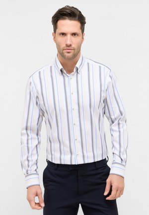 Man wearing a long-sleeve button-up shirt with vertical blue, gray, and white stripes, paired with dark pants, standing against a plain background.