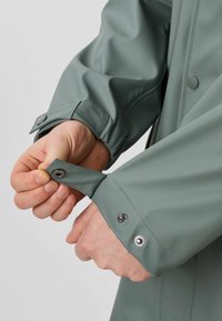 Mani che sistemano i polsini con bottoni automatici su una manica di una giacca impermeabile verde con una finitura opaca liscia.