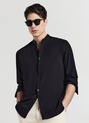 Chemise noire à boutons avec col, en tissu texturé et coupe décontractée. Portée avec des lunettes de soleil et un pantalon couleur crème.