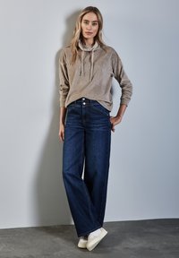 Beige hoodie med snoddar, kombinerad med vida mörkblå jeans och vita sneakers, framför en enfärgad grå bakgrund.