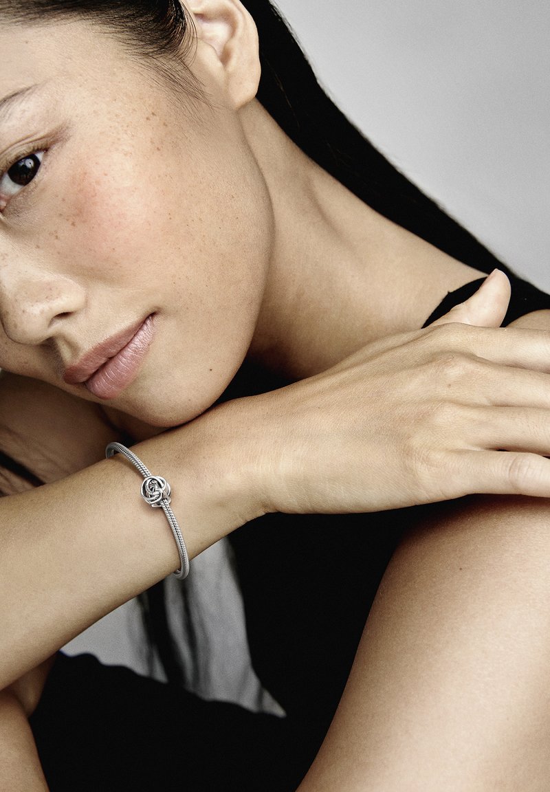 Bracciale d'argento con un design intrecciato e un dettaglio decorativo a nodo, indossato su un polso. La pelle del modello presenta una texture naturale con lentiggini.