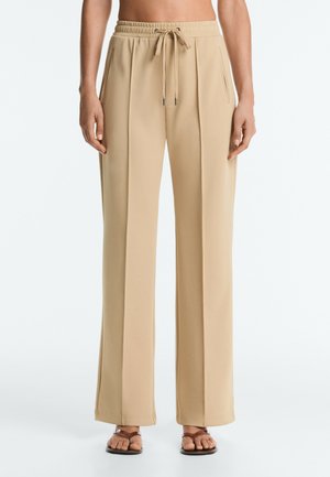 Pantalon beige à jambes larges avec pli devant, taille élastique et cordon, porté avec des sandales plates marron sur une personne debout.