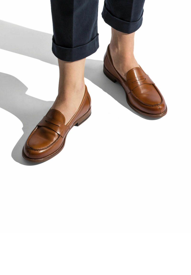 Mocasines de cuero marrón con acabado liso y diseño cosido. Punta redonda y tacón bajo. Usados con pantalones azul marino remangados.