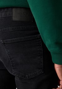 Crne traperice s pravokutnom stražnjom džepnom, na kojoj se nalazi teksturirana kožna oznaka s natpisom "Marc O'Polo DENIM." Iznad se vidi zeleni materijal.