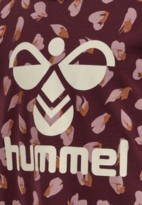 Weißes Hummel-Logo und Text auf bordeauxrotem Stoff mit pinkem und braunem Blumenmuster im Hintergrund.