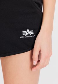 Čierne bavlnené šortky s rebrovanou textúrou, s bielym logom "ALPHA INDUSTRIES" a trojuholníkovou grafikou po boku.