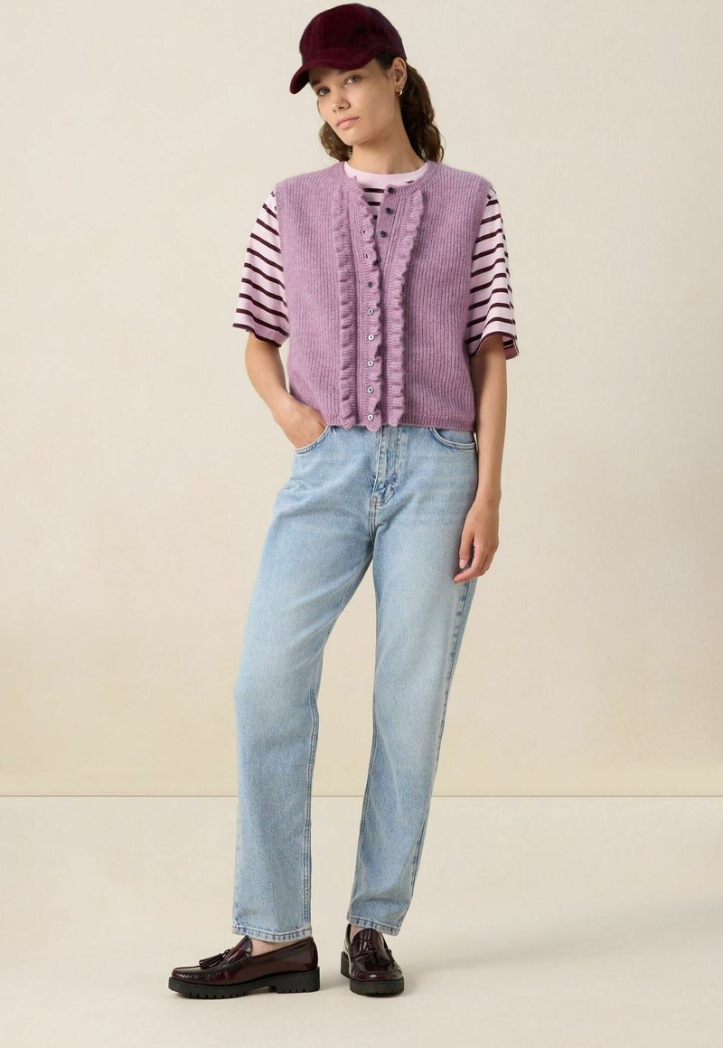 Femme se tenant debout avec une main dans la poche, portant une casquette bordeaux, une chemise rayée, un gilet en maille boutoné lavande, un jean bleu clair et des mocassins foncés.