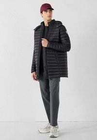 Giubbotto a vento nero con cuciture orizzontali e cappuccio, indossato sopra una maglietta nera, abbinato a pantaloni grigi e sneaker bianche, con un berretto bordeaux.