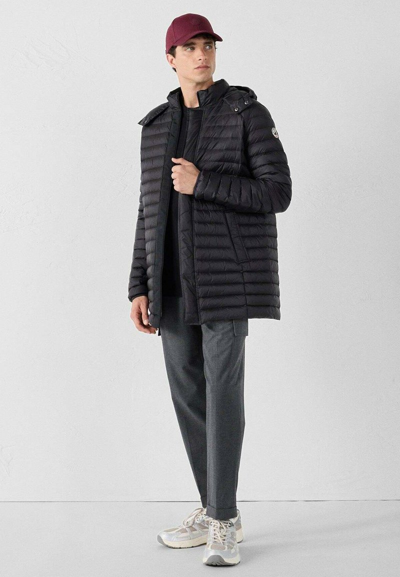 Giubbotto a vento nero con cuciture orizzontali e cappuccio, indossato sopra una maglietta nera, abbinato a pantaloni grigi e sneaker bianche, con un berretto bordeaux.