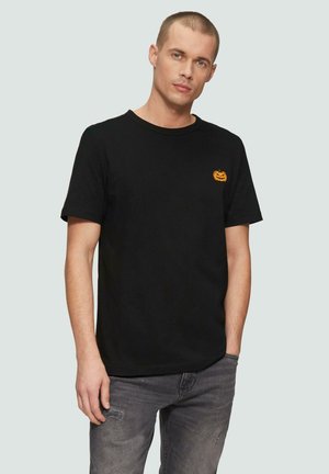 Schwarzes Baumwoll-T-Shirt mit einer kleinen orangefarbenen Kürbisstickerei auf der linken Brustseite, kombiniert mit grauen, stark getragenen Jeans.