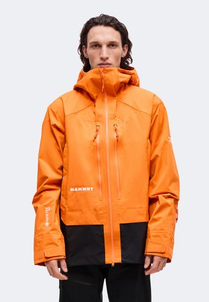 Un homme porte une veste imperméable orange Mammut à col montant, avec un empiècement inférieur noir et des poches à l’avant, sur un fond uni.