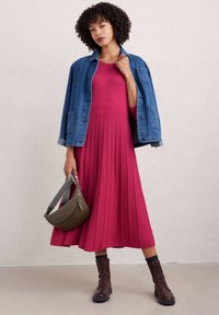 Abito plissettato fucsia con maniche corte, abbinato a un giubbotto di jeans e una borsa verde oliva. Stivali marroni con lacci completano il look.