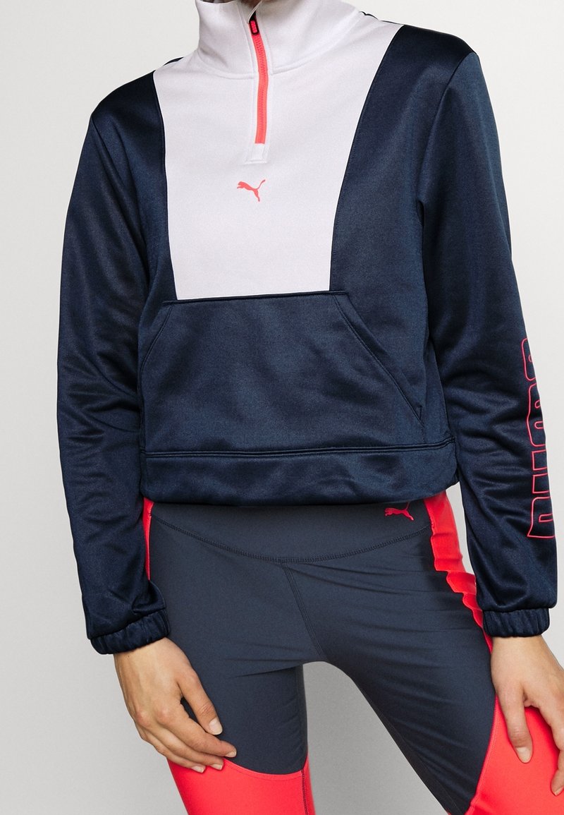 Femme portant un sweat zippé Puma bleu marine et blanc avec une poche devant, associé à des leggings de sport bleu marine et rouge assortis.