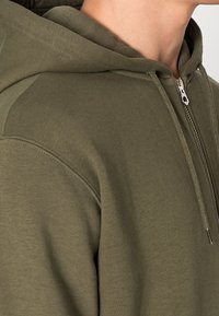 Nahaufnahme einer Person, die einen olivgrünen Hoodie mit einem Reißverschluss vorne, Kordeln und sichtbarer Kapuze trägt, wobei die obere Schulter und der Hals zu sehen sind.