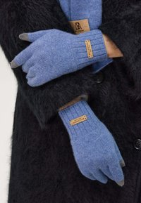 Gants en tricot bleus avec des bouts gris et des poignets côtelés, arborant une étiquette. Ils sont portés par-dessus un manteau noir et duveteux.