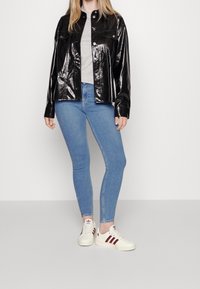 Chaqueta negra, brillante y de gran tamaño con botones a presión, llevada sobre una camisa gris y unos pantalones vaqueros ajustados de color azul claro; combinada con zapatillas blancas con franjas rojas.