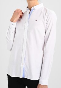 Camisa blanca de manga larga con cuello tipo botón, que presenta un forro interior azul y un pequeño logo bordado en el pecho. Textura suave.